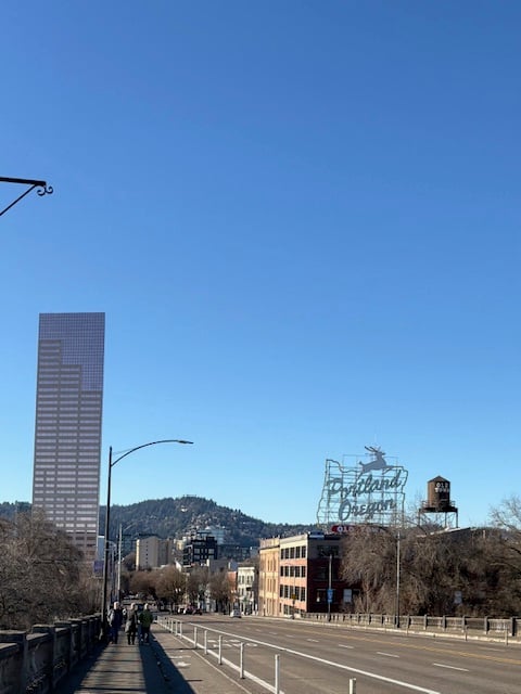 On the left the pink skyscraper from the previous photo, in the center in the distance a tree-covered hill, on the right an unlit neon sign (it's daytime) in the shape of the state of Oregon. It says Portland, Oregon. A stylized white stag leaps across the top. 