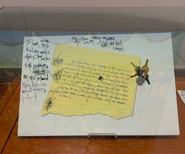 A set of keys and a torn sheet from a yellow legal pad. Text says On this chain is a key that I no longer use but still carry. I look at the keys and all the words that I have written and the scabbling doodling on this yellowed paper that reminds of old newspapers and headlines.