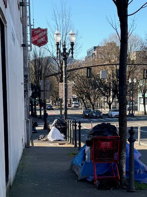 Two tents pitched on the sidewalk under a Salvation Army sign. One is neat, with a red shopping cart parked next to it, a black plastic bag in the cart. The other has more debris scattered around the perimeter.