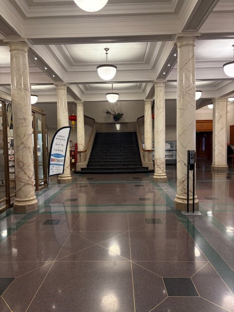 Library lobby with brown marble floors, cream-colored marble columns, and a sweeping central staircase. Molded ceiling with drop light fixtures. 