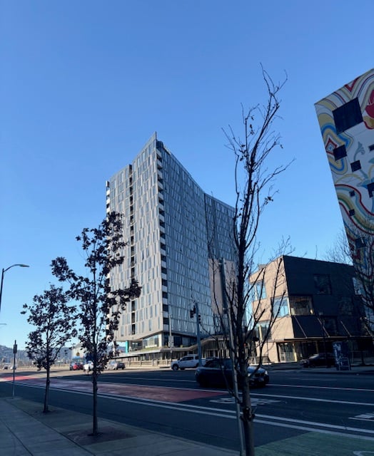 A tall, dark, angular building seen from across the street. There is a small, squat brown building, also of modern design to its right. To its left is only blue sky because it sits right by the river.