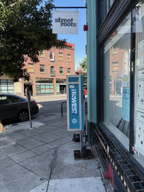 Small sign that says Street Roots hanging over a functional payphone on a city street.